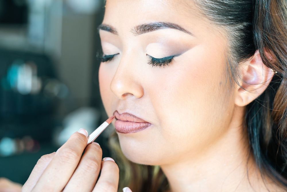 A makeup artist applies lip gloss to a young woman with elegant eye makeup and long lashes in a beauty salon.