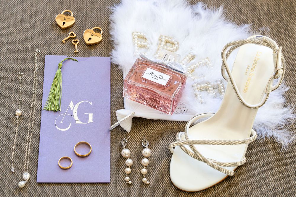 Elegant wedding flat lay with gold rings, Dior perfume, pearl earrings, white heels, lock charms, and monogram stationery.