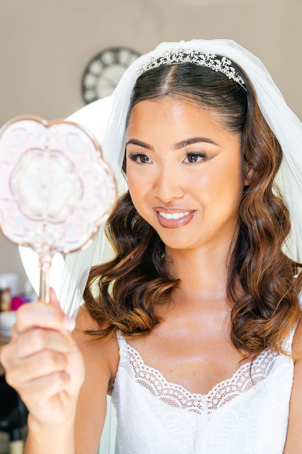 Smiling bride in white lace dress and tiara holds decorative hand mirror on her wedding day.