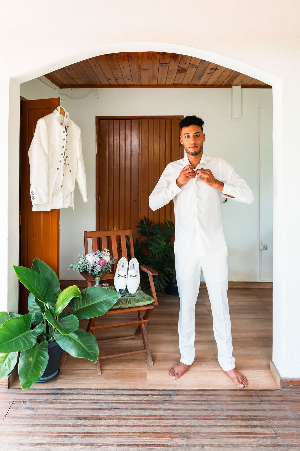 Young man in white outfit buttoning shirt barefoot in elegant room with wooden chair, plants, and white shoes nearby.