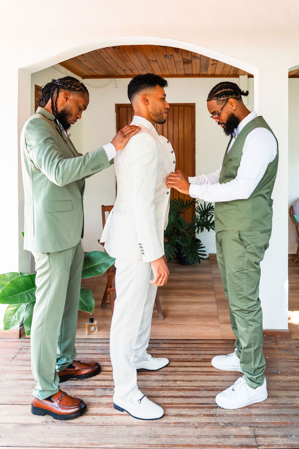 Groom in white suit being helped by two groomsmen in sage green suits during wedding preparations indoors.