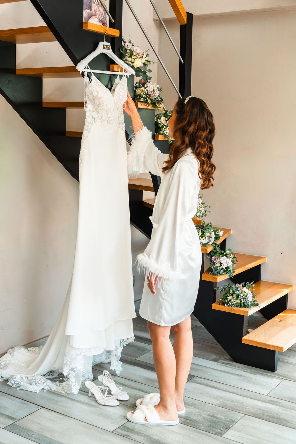 Bride in white robe admiring her wedding dress hanging on staircase, with heels and floral decorations nearby.