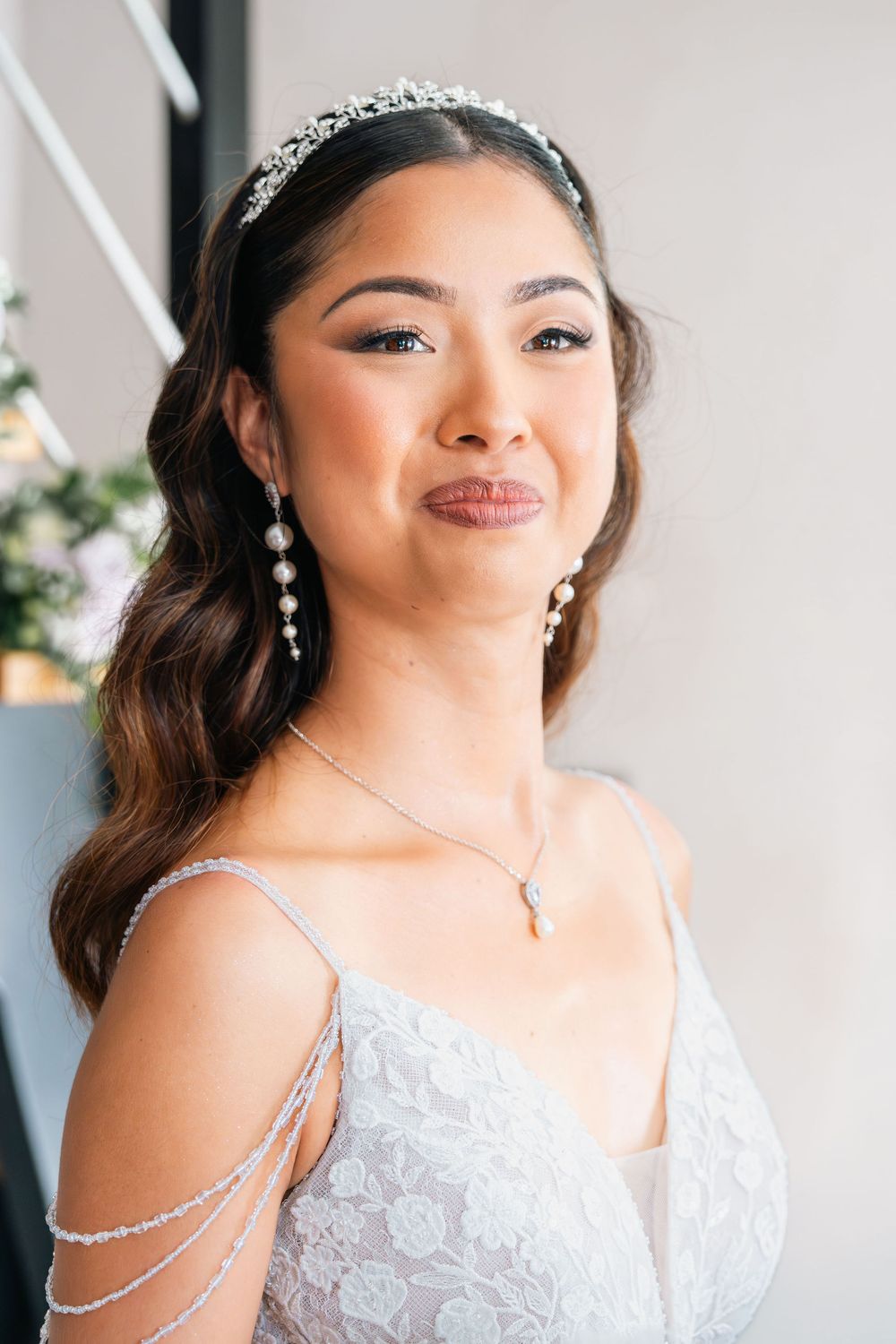 Bride wearing crystal tiara, pearl drop earrings, and lace gown with delicate shoulder jewelry, smiling softly.