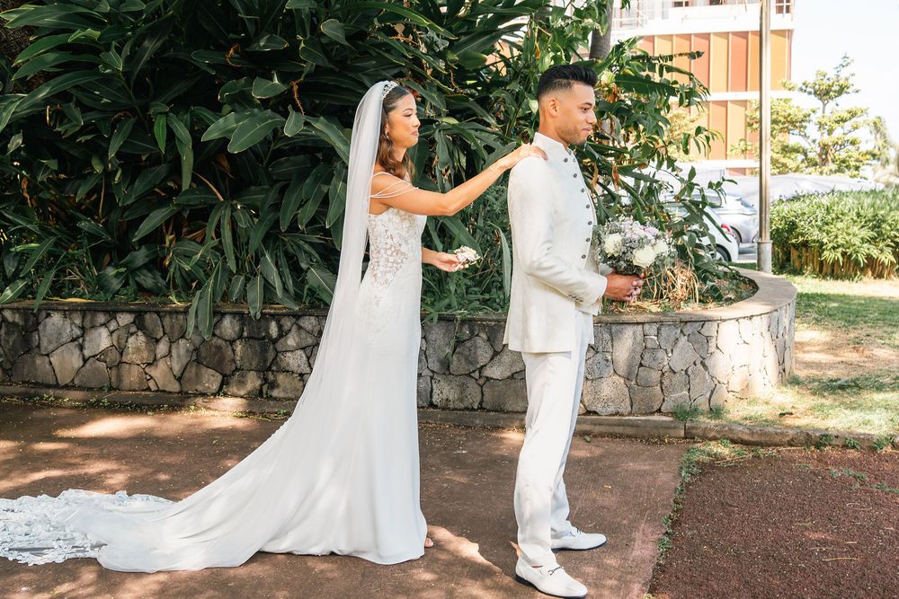 Bride in white gown touches groom's shoulder during first look moment outdoors near stone wall and tropical greenery.