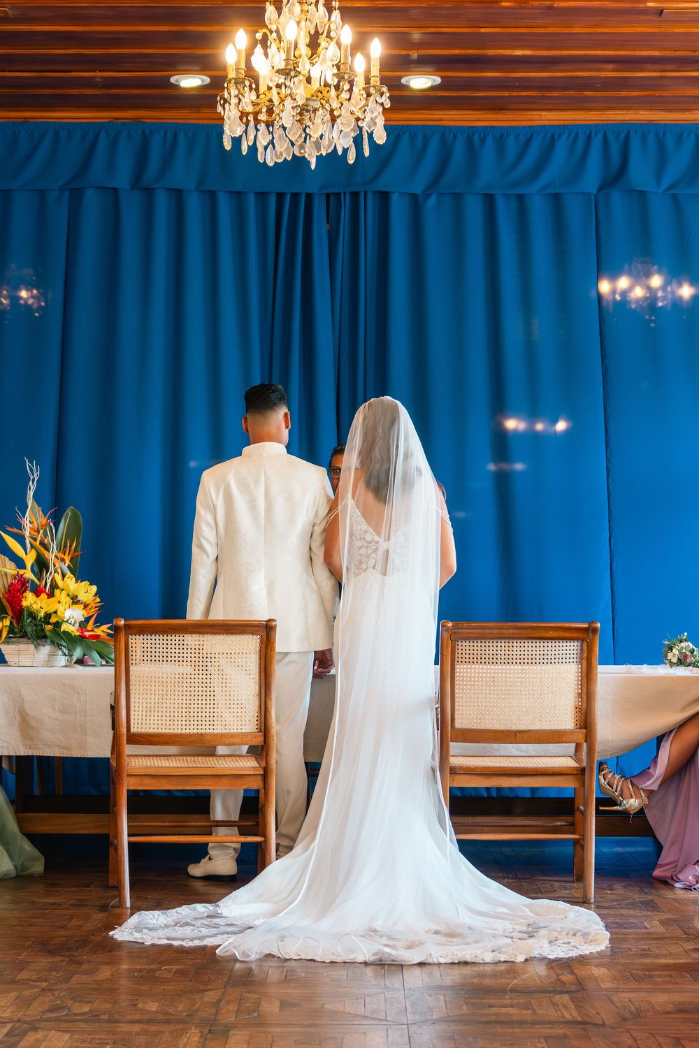 Bride and groom standing at wedding altar with blue curtain backdrop and crystal chandelier overhead.