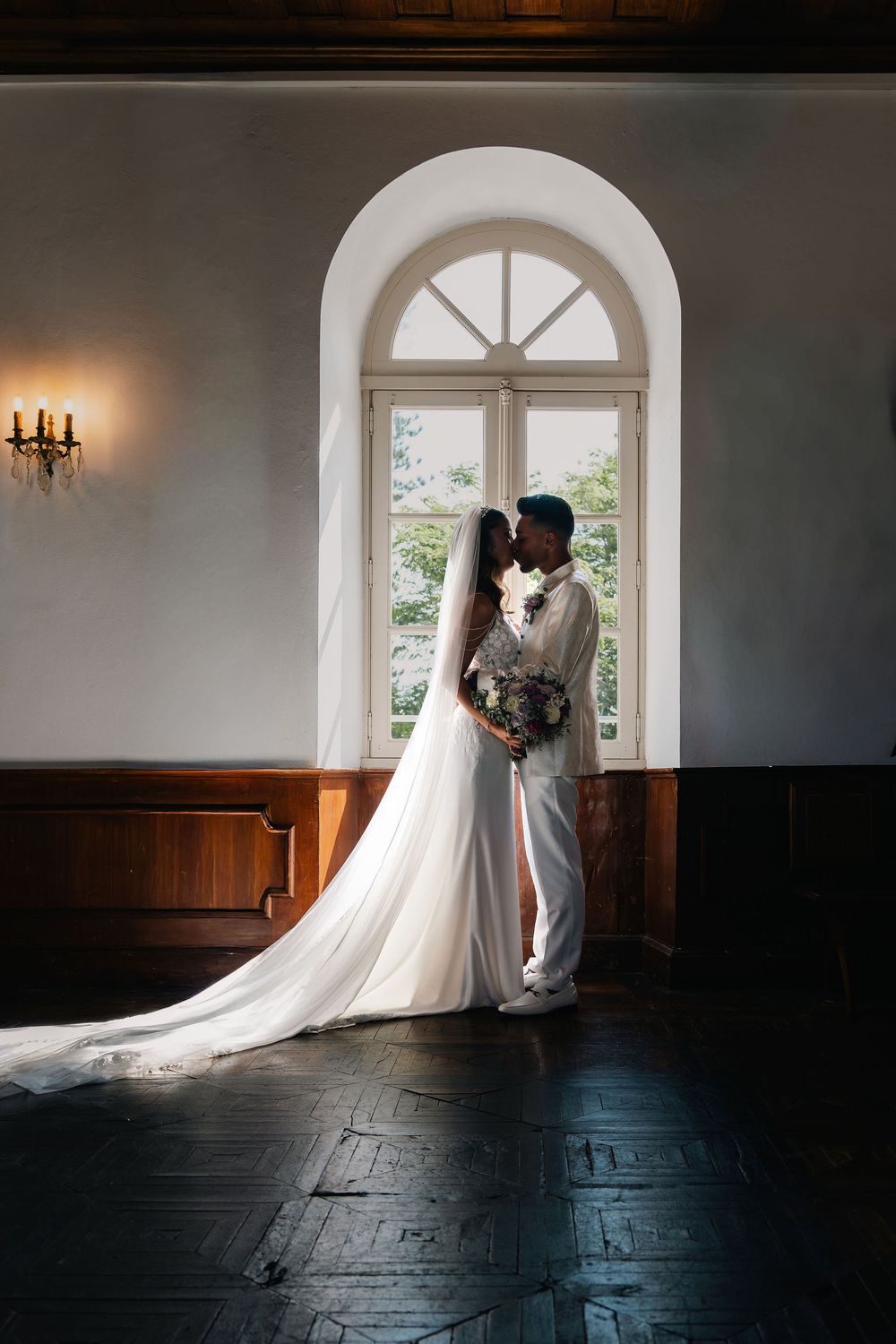 Bride and groom kissing in front of arched window, bride in elegant white gown with long train in a classic venue.