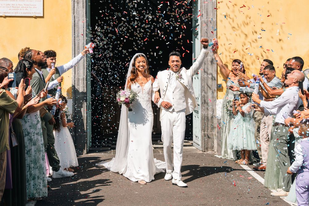 Newlywed couple exits church amid colorful confetti shower, guests celebrating joyful wedding ceremony outdoors.