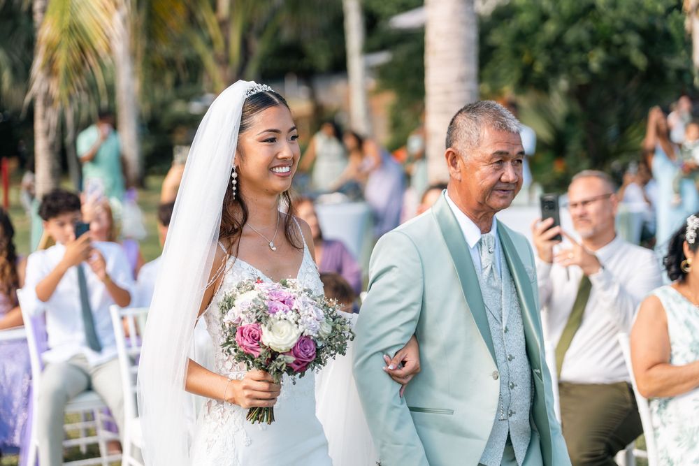Bride in white gown and veil walks down outdoor aisle with older man in mint suit, holding colorful bouquet.