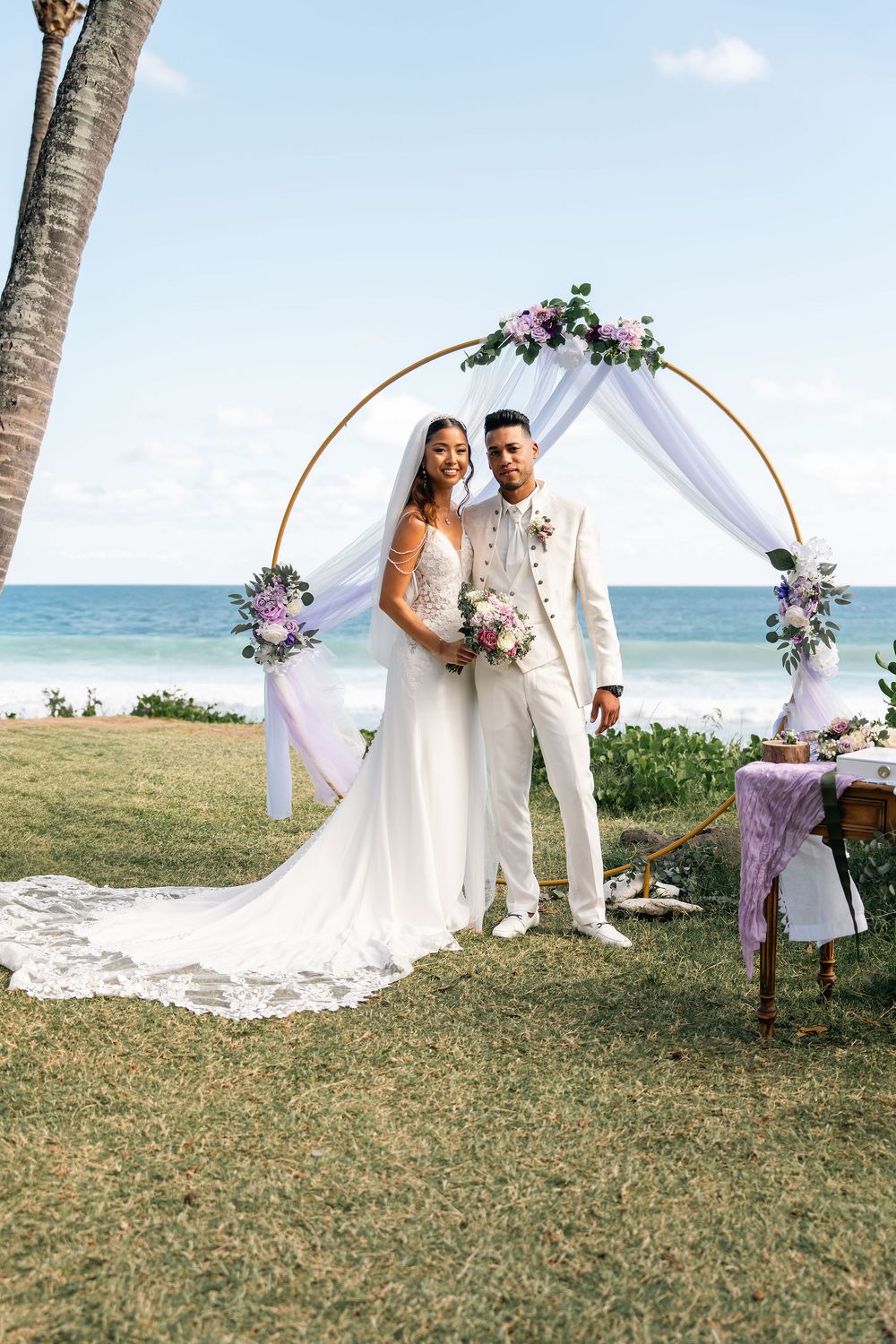 Bride and groom posing under a circular floral arch with ocean backdrop at a beachside wedding ceremony.