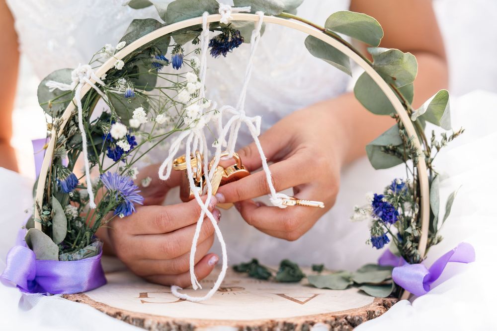 Bride holding wedding rings on a decorative hoop with eucalyptus, blue flowers, and white ribbon at a wedding ceremony.