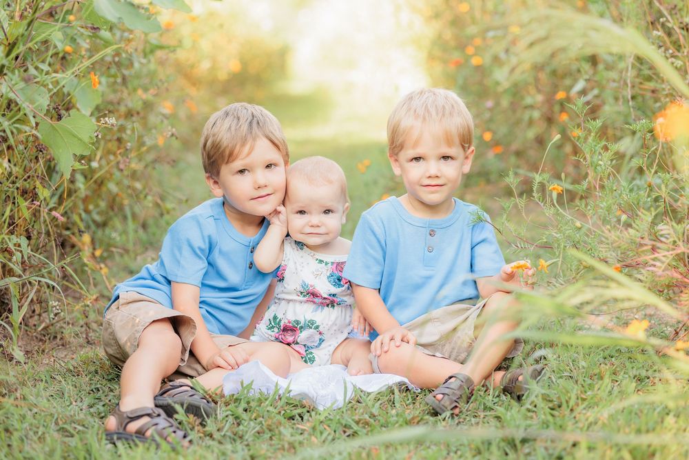 Three young siblings sit together in a lush green field, two boys in blue shirts flanking a baby girl in a floral dress.
