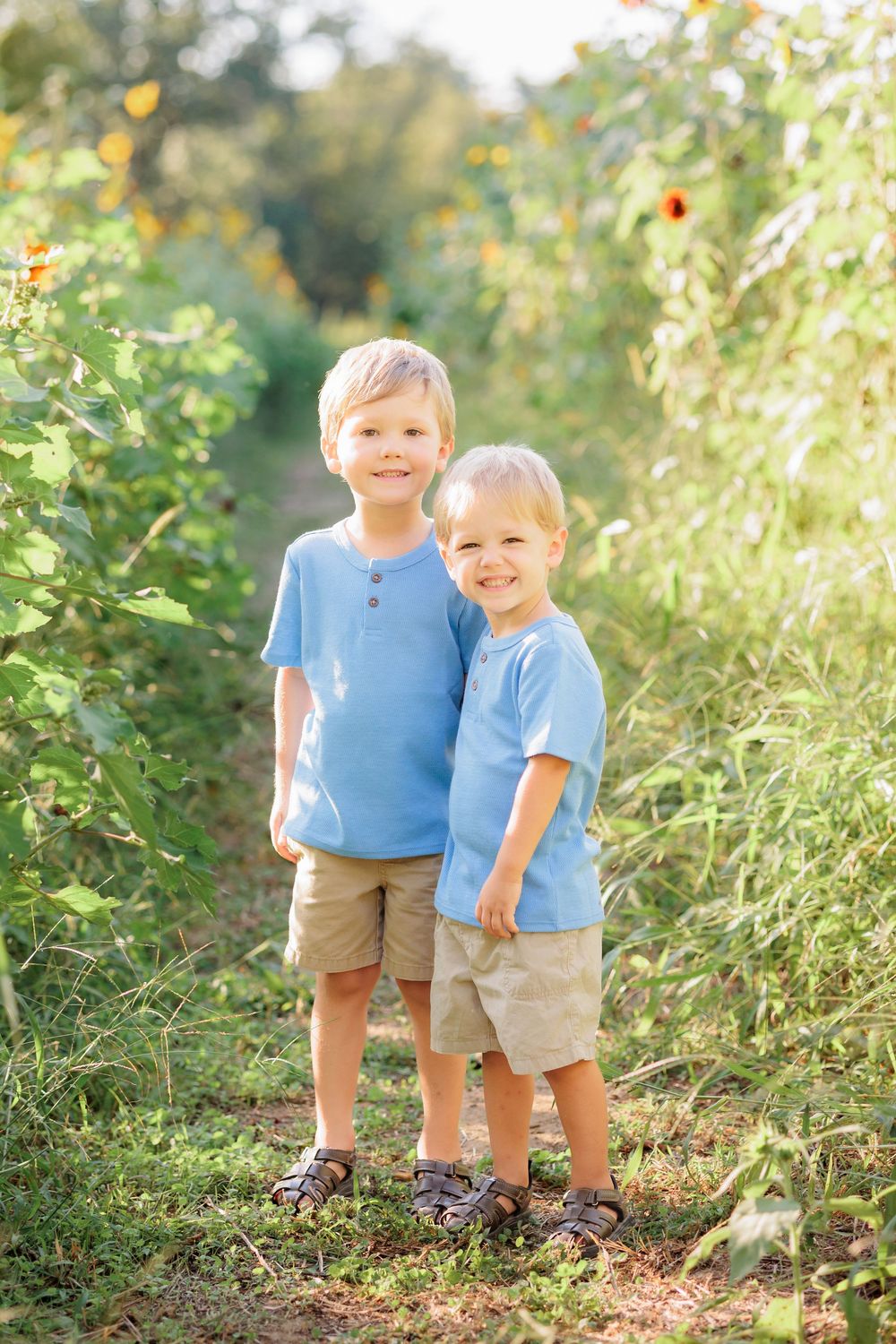 Two young brothers in matching blue shirts and tan shorts smile together on a sunny garden path.