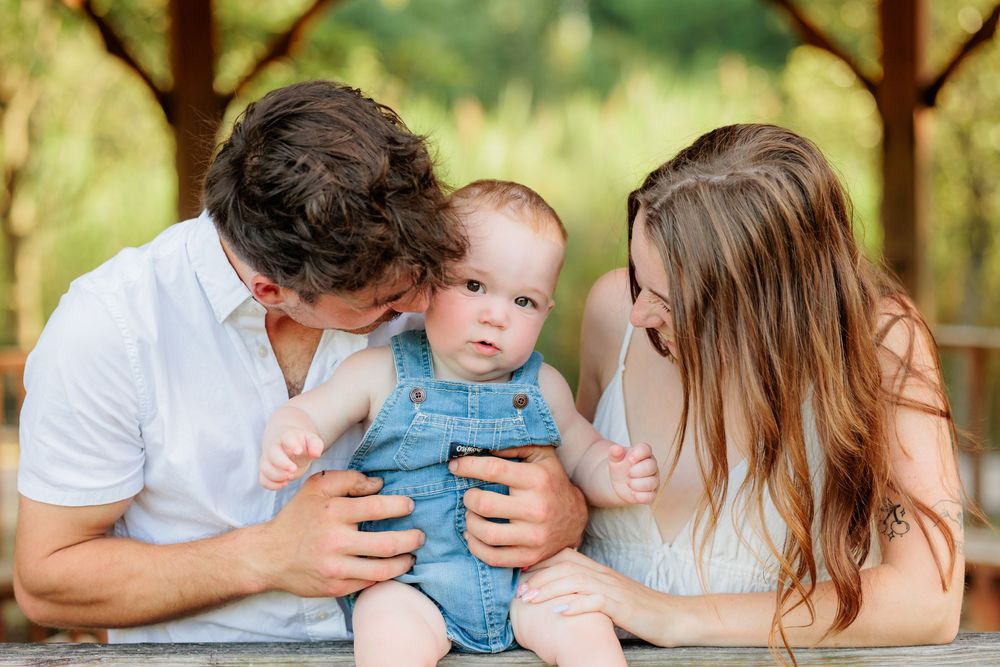 A young family with parents kissing and smiling at their baby boy in denim overalls outdoors.