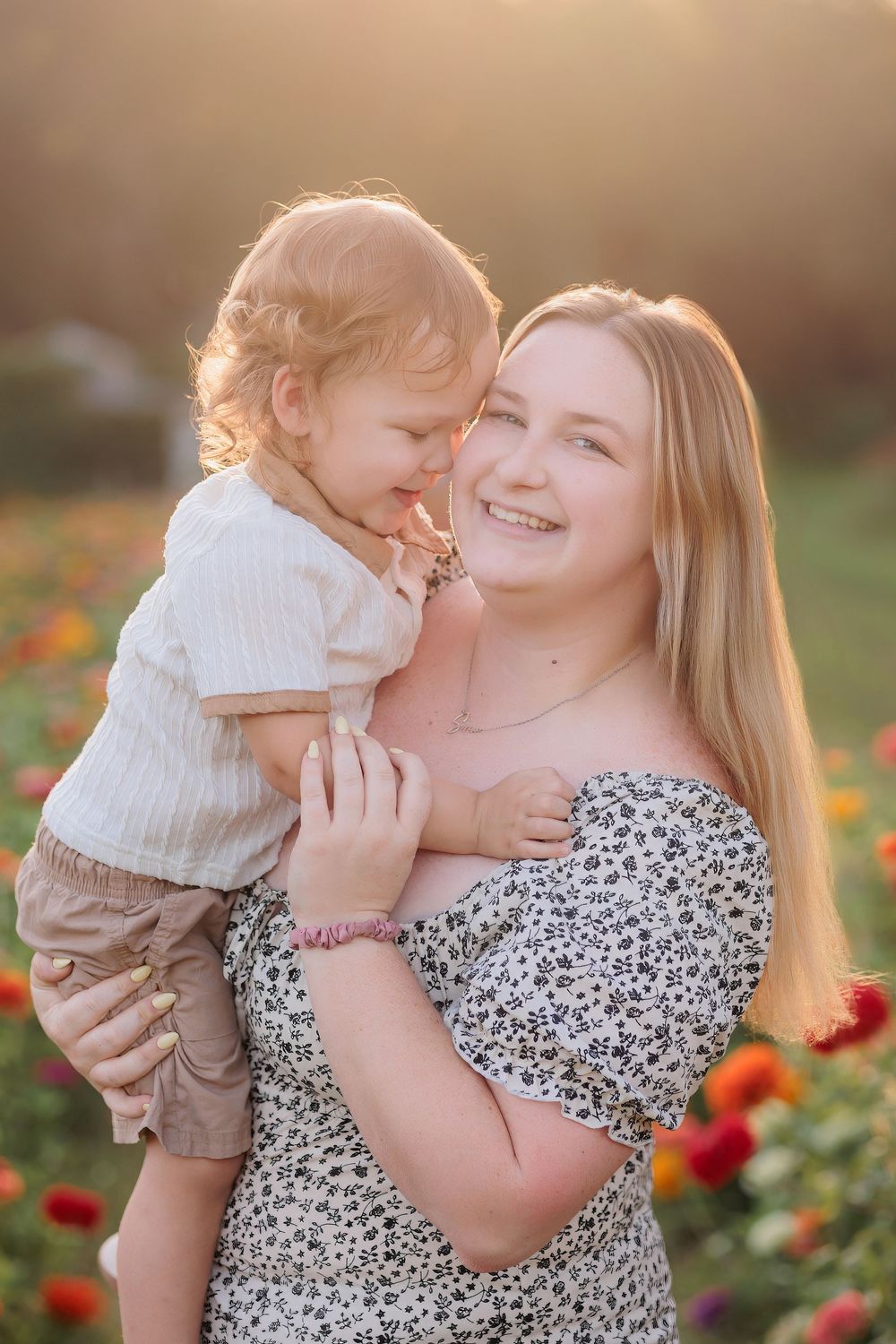 A smiling mother holds her curly-haired toddler in a sunlit flower field at golden hour.