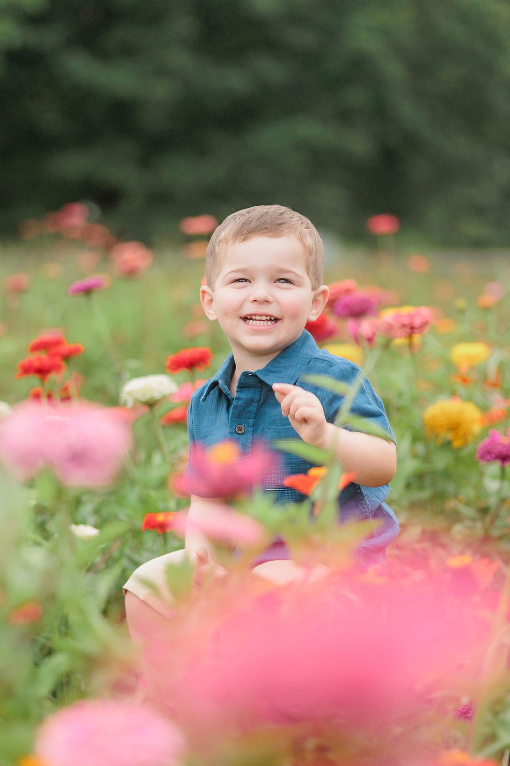 A cheerful toddler boy in a blue shirt smiles brightly while sitting among colorful zinnia flowers in a garden.
