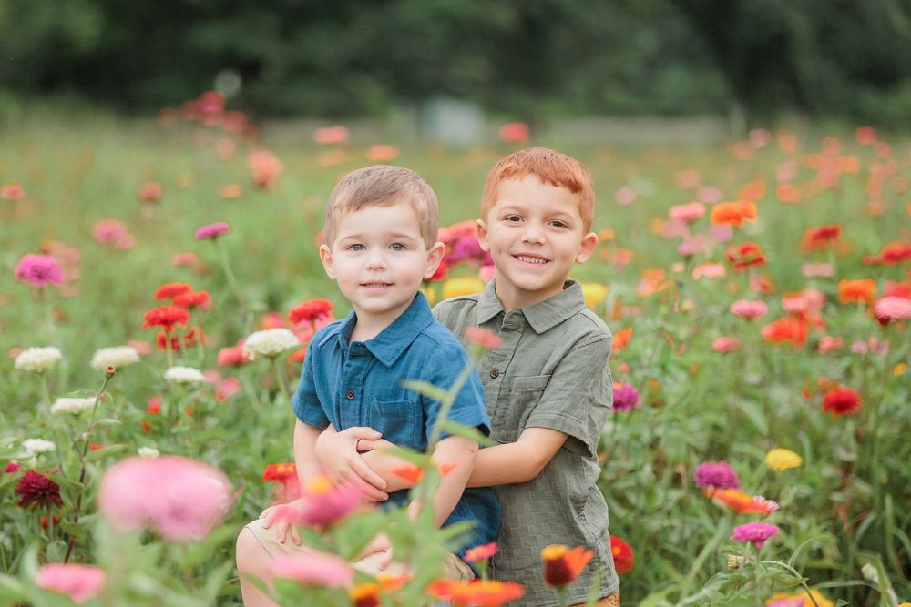 Two young red-haired boys smiling in a vibrant flower field filled with colorful zinnias in pink, red, and orange.