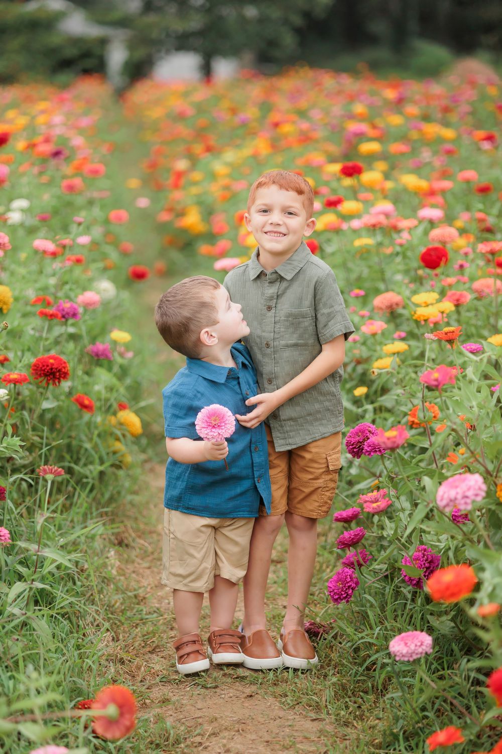 Two young red-haired boys smile together in a vibrant flower field filled with colorful dahlias and zinnias.