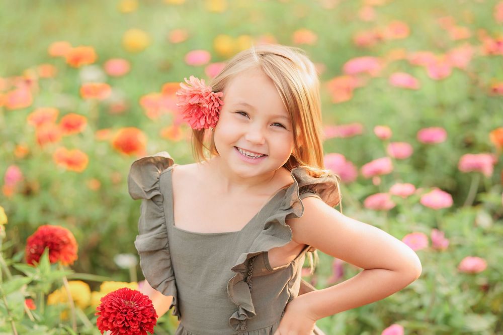 Smiling young girl with a pink flower in her hair poses in a vibrant garden full of colorful zinnia blooms.