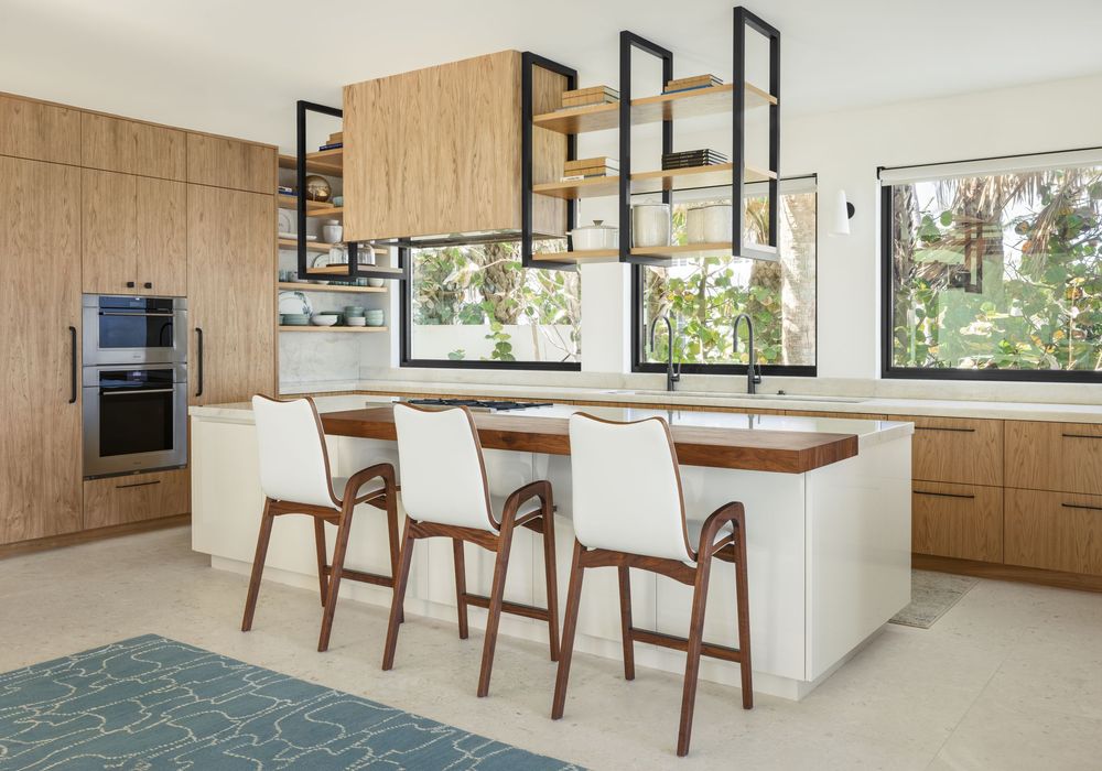Modern kitchen with warm wood cabinets, white island, open metal shelving, and white upholstered bar stools.