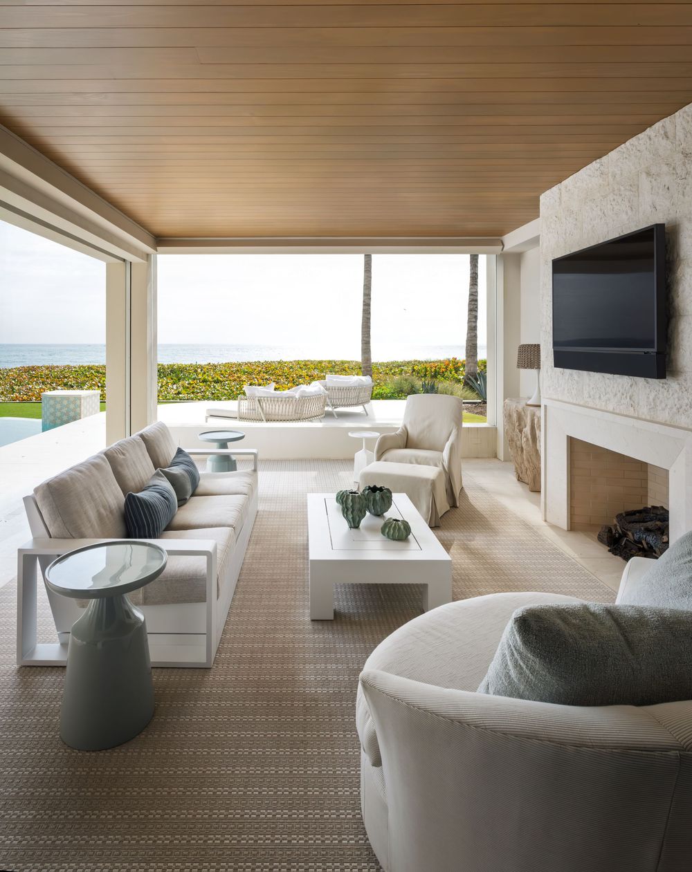 Luxurious covered oceanfront patio with neutral sectional, white coffee table, stone fireplace, and panoramic sea views.