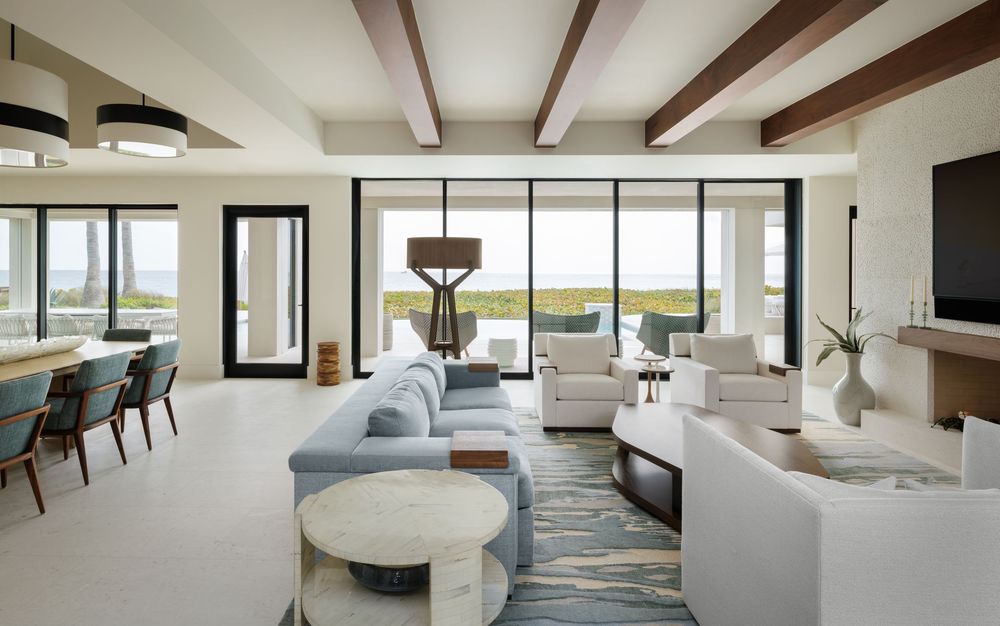 Bright coastal living room with exposed wood beams, blue sectional, white curved sofa, and floor-to-ceiling windows.