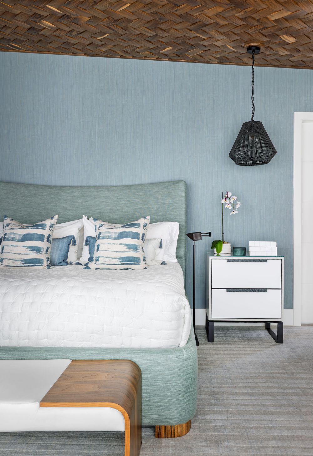 Serene blue bedroom with upholstered headboard, herringbone wood ceiling, white bedding, and black pendant light.