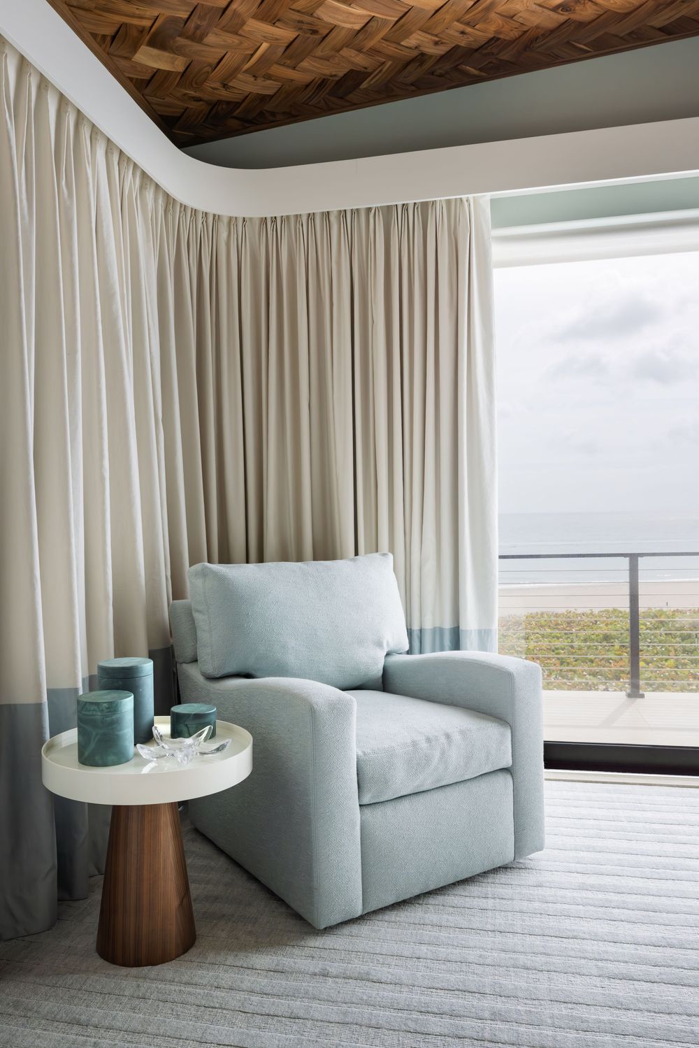 Coastal reading nook with light blue armchair, round pedestal side table, floor-length curtains, and ocean view balcony.