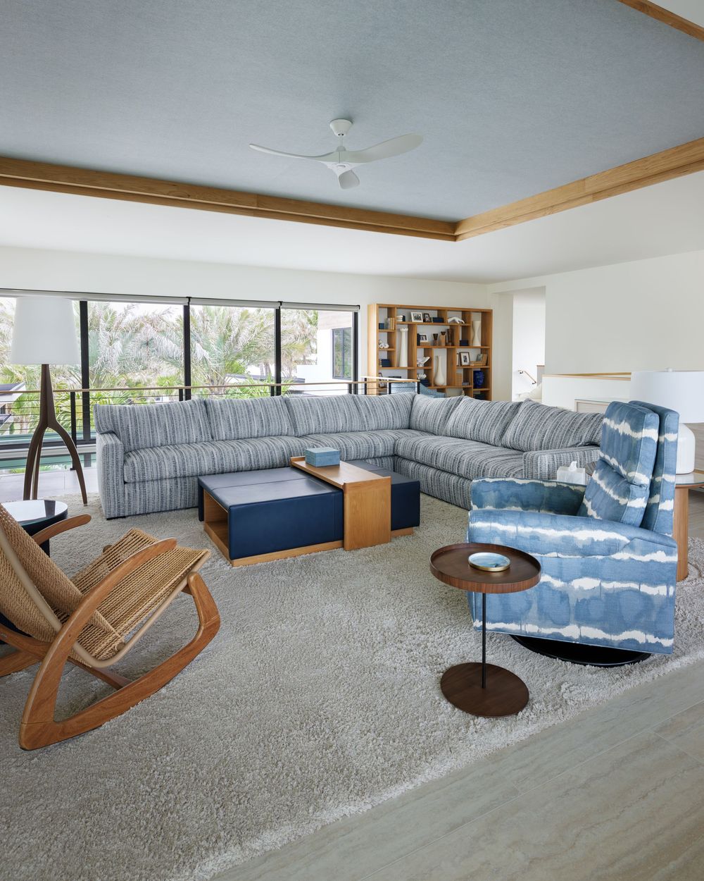 Spacious living room with striped sectional sofa, blue accent chair, rocking chair, and panoramic windows overlooking palms.