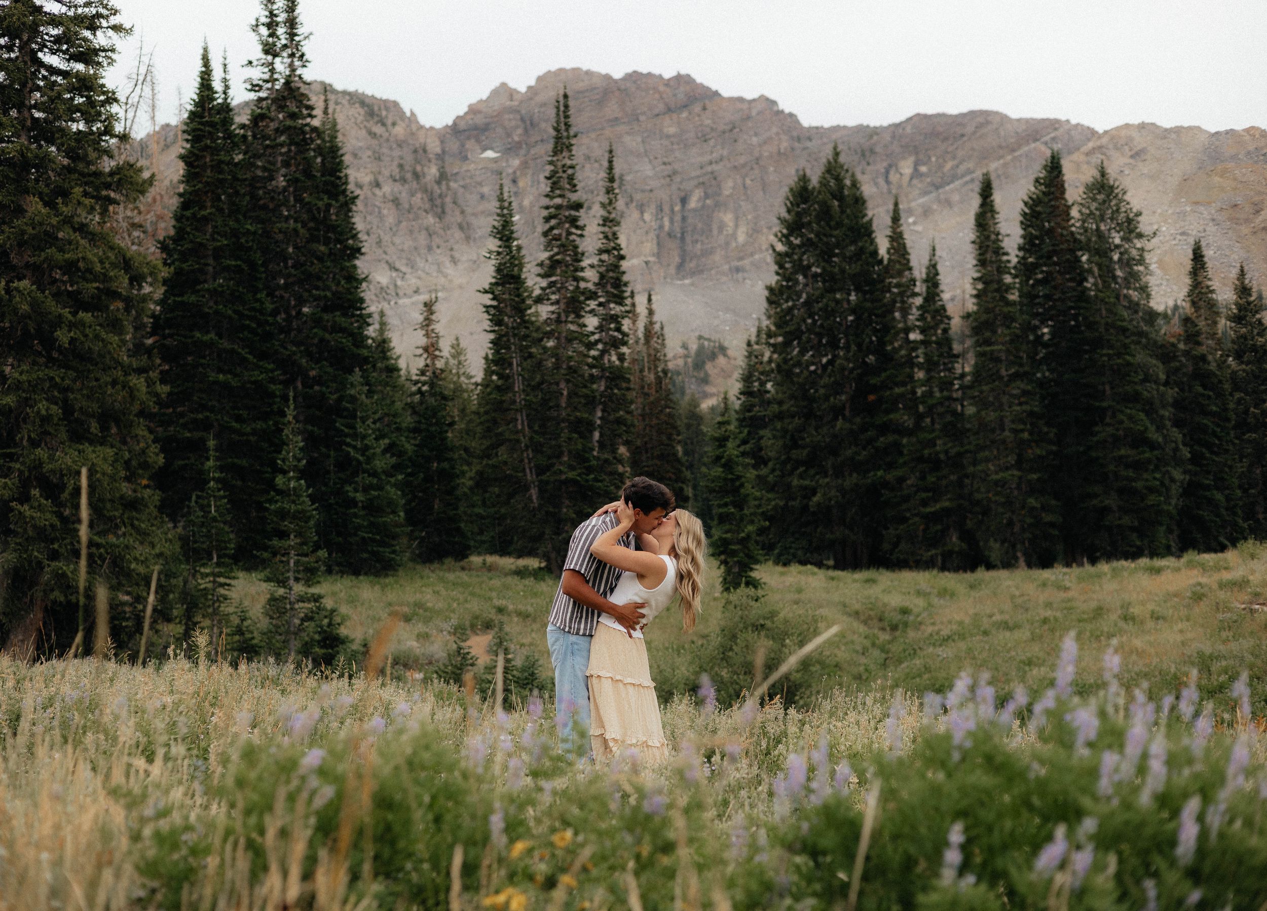 Couple embracing in a wildflower meadow with tall pine trees and rocky mountains in the background.