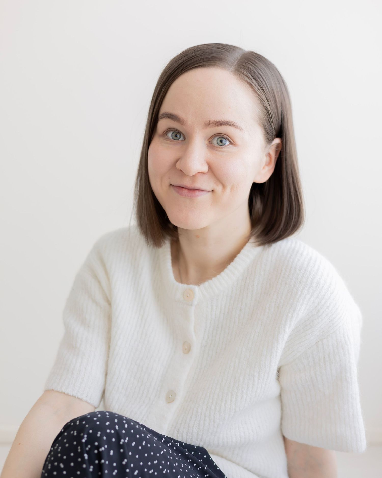 Young woman with short brown hair wearing a white knit cardigan, smiling softly against a light background.