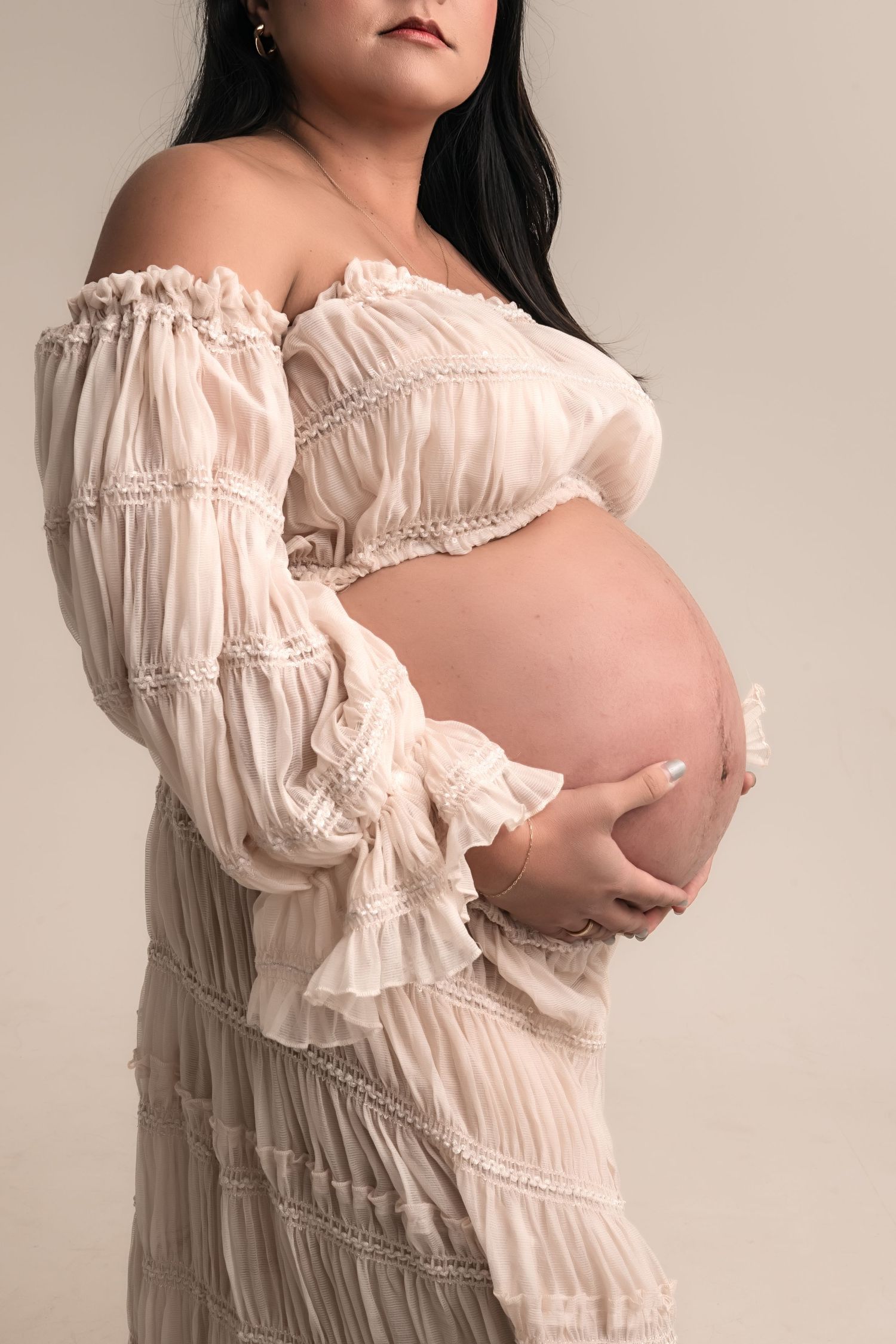 Pregnant woman in a flowing off-shoulder cream ruffle dress cradling her baby bump against a neutral background.