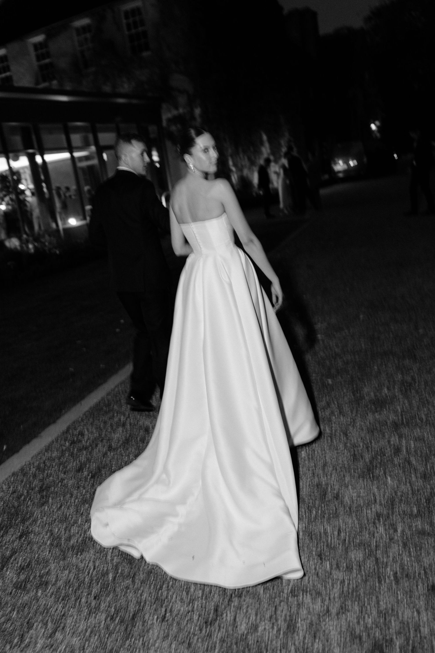 Black and white photo of bride in strapless ball gown with long train walking with groom in tuxedo at night.