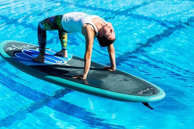 Person practicing yoga on a paddleboard in a bright blue swimming pool, performing a backbend pose.