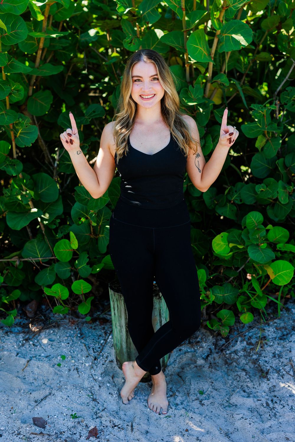 A smiling woman in a black outfit points upward with both hands while standing barefoot in front of lush green foliage.