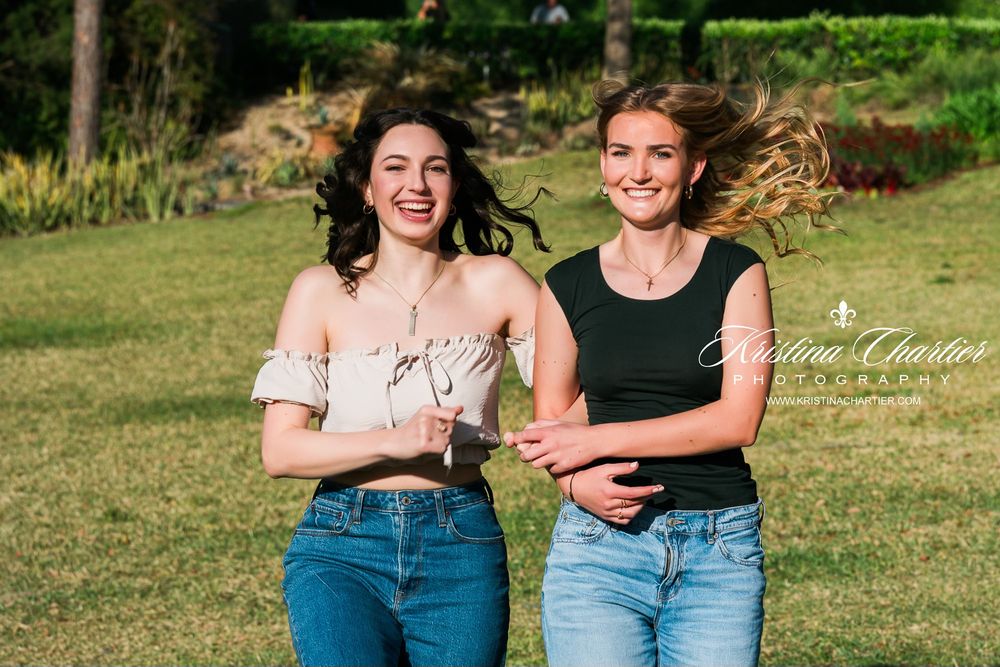 Two young women laughing and running together on green grass in a sunny outdoor park setting.
