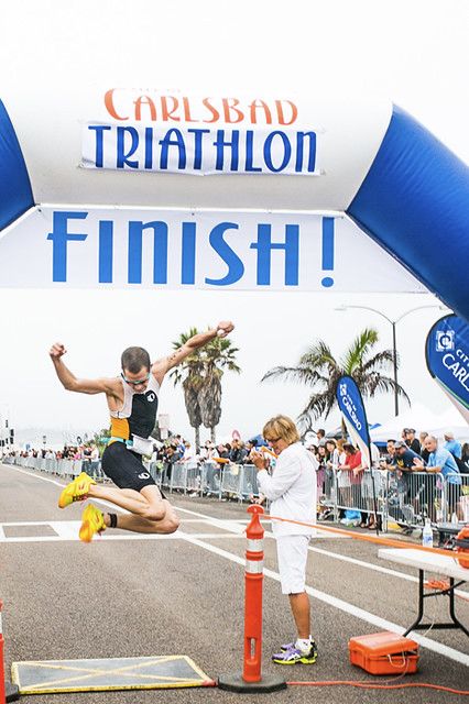 Athlete leaping triumphantly across the finish line at the Carlsbad Triathlon race event.