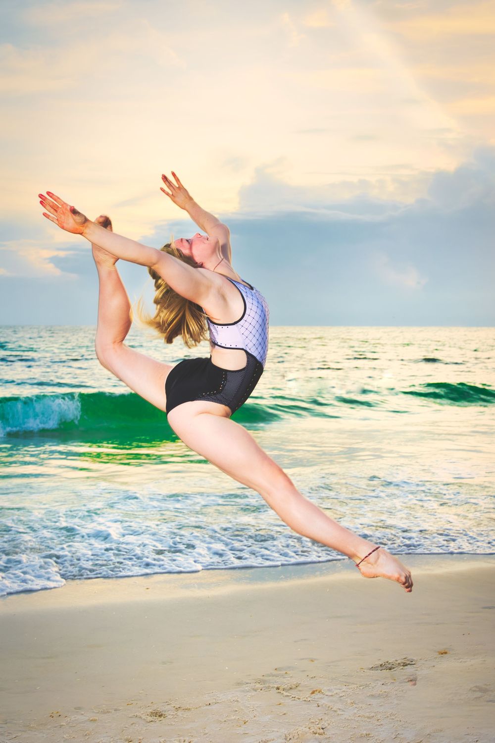 A gymnast leaps gracefully on a beach at sunset, performing a split jump in a black and white swimsuit.