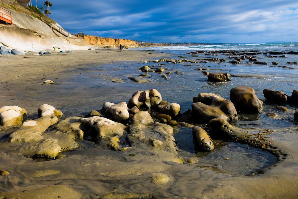 Rocky shoreline with moss-covered boulders along a sandy beach with dramatic cliffs and blue sky.