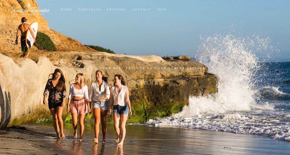 Four women walk along a sunny beach shoreline as waves crash on rocks and a surfer walks in the background.