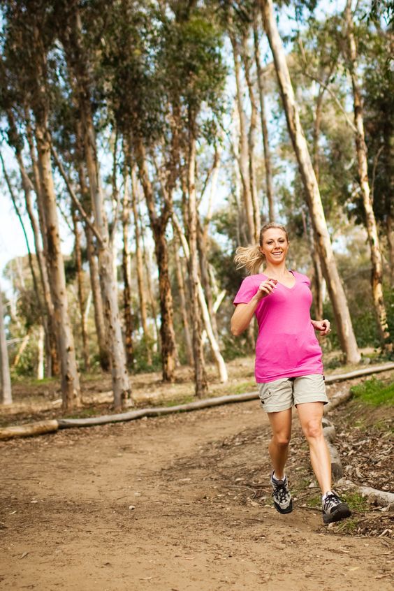 A smiling blonde woman in a pink shirt jogs on a dirt trail through a sunny eucalyptus forest.