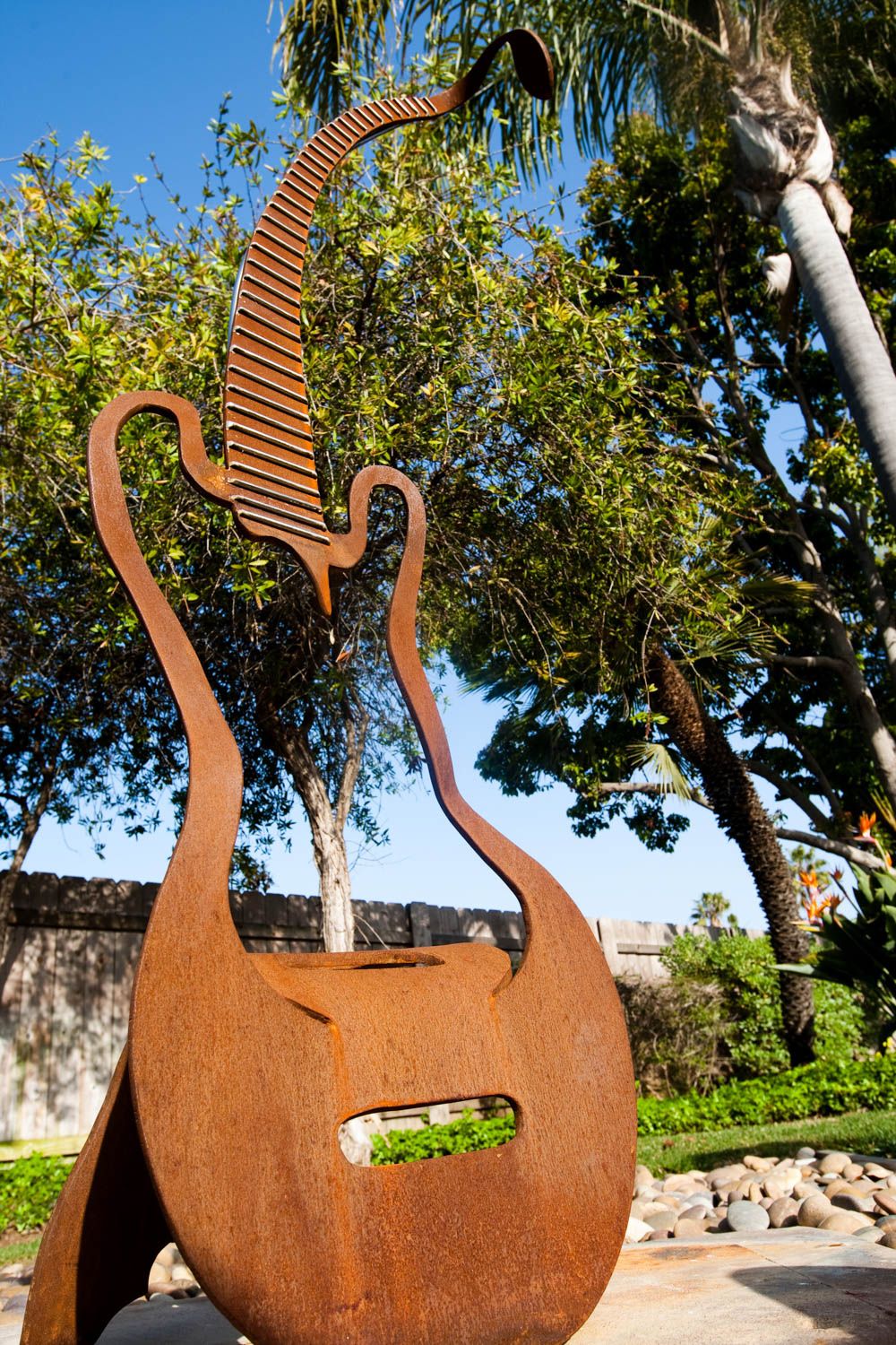Rusty metal lyre sculpture in an outdoor garden setting with palm trees and blue sky backdrop.