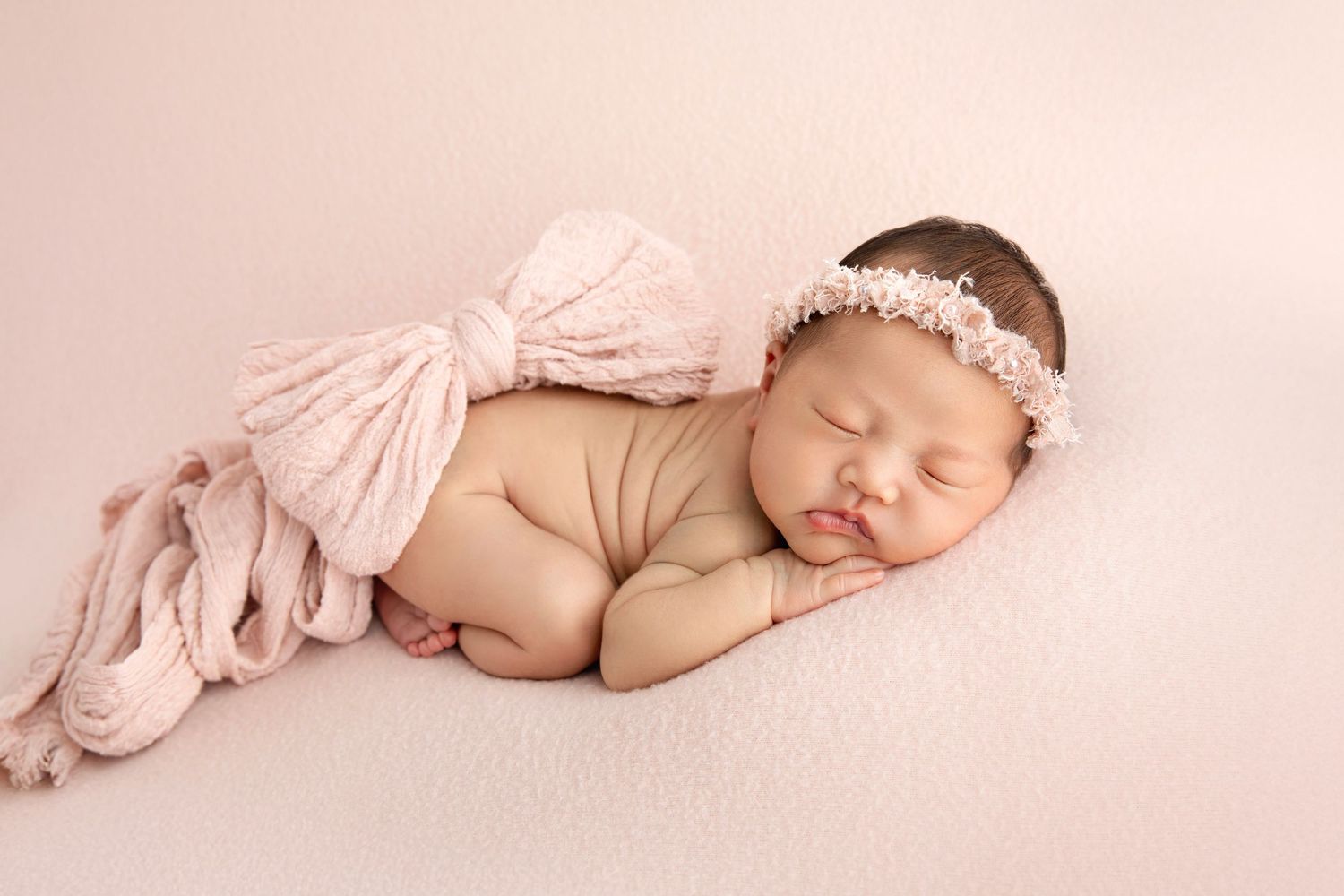 Sleeping newborn baby girl in pink wrap and floral headband on soft pink blanket in studio newborn photo.