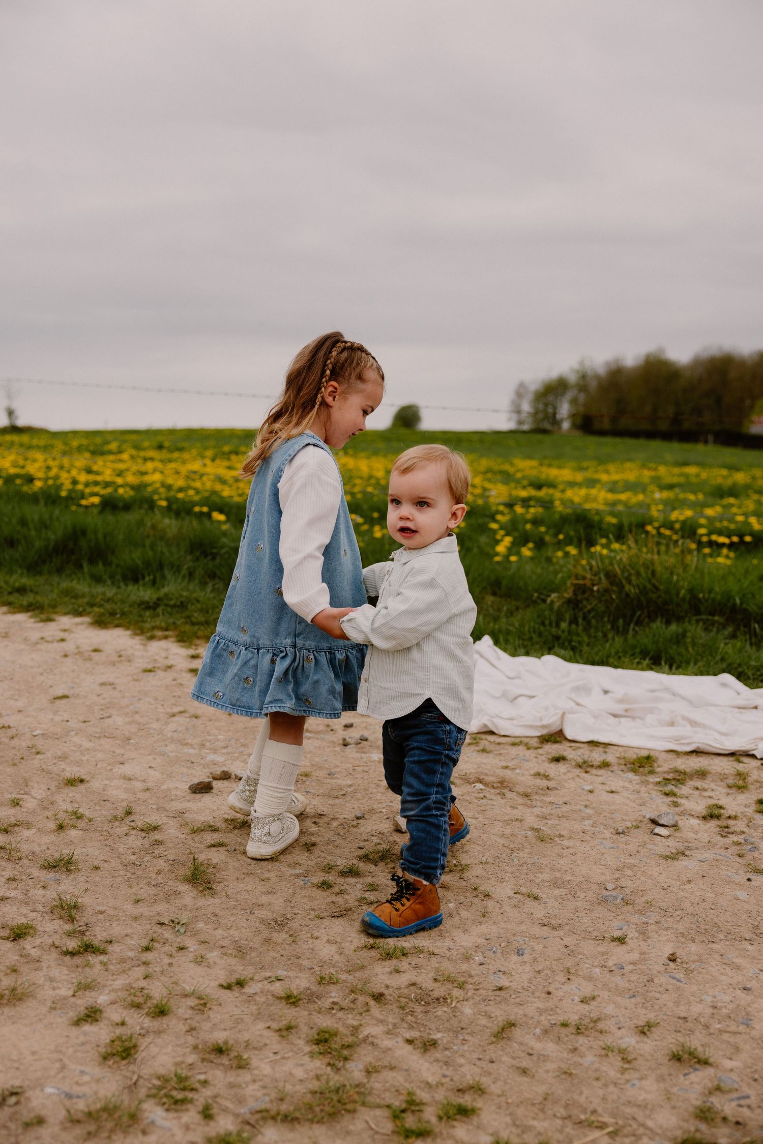 Two young siblings holding hands on a dirt path beside a yellow flower field.