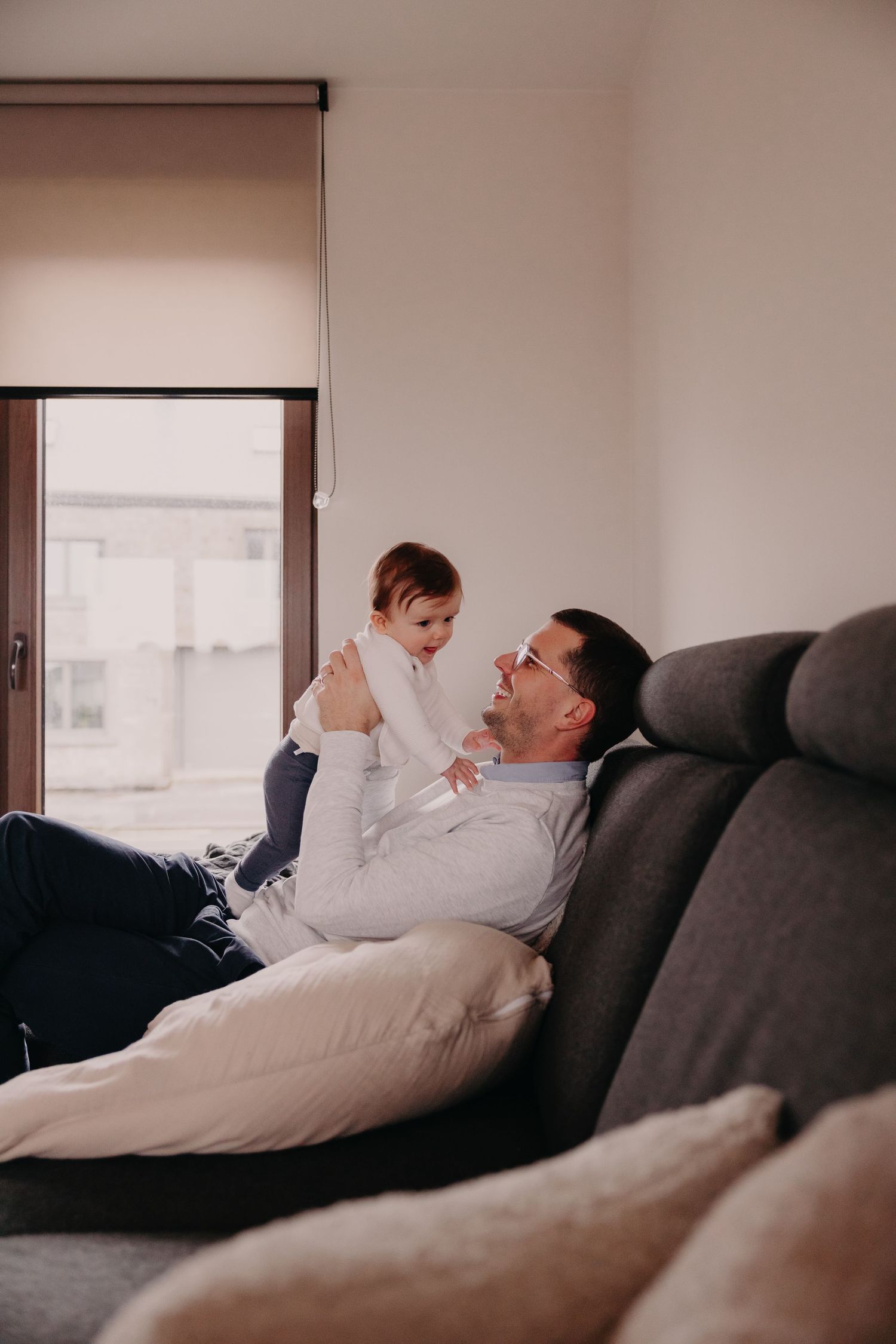 Father lying on gray sofa lifting and playing with his laughing toddler in a bright living room.