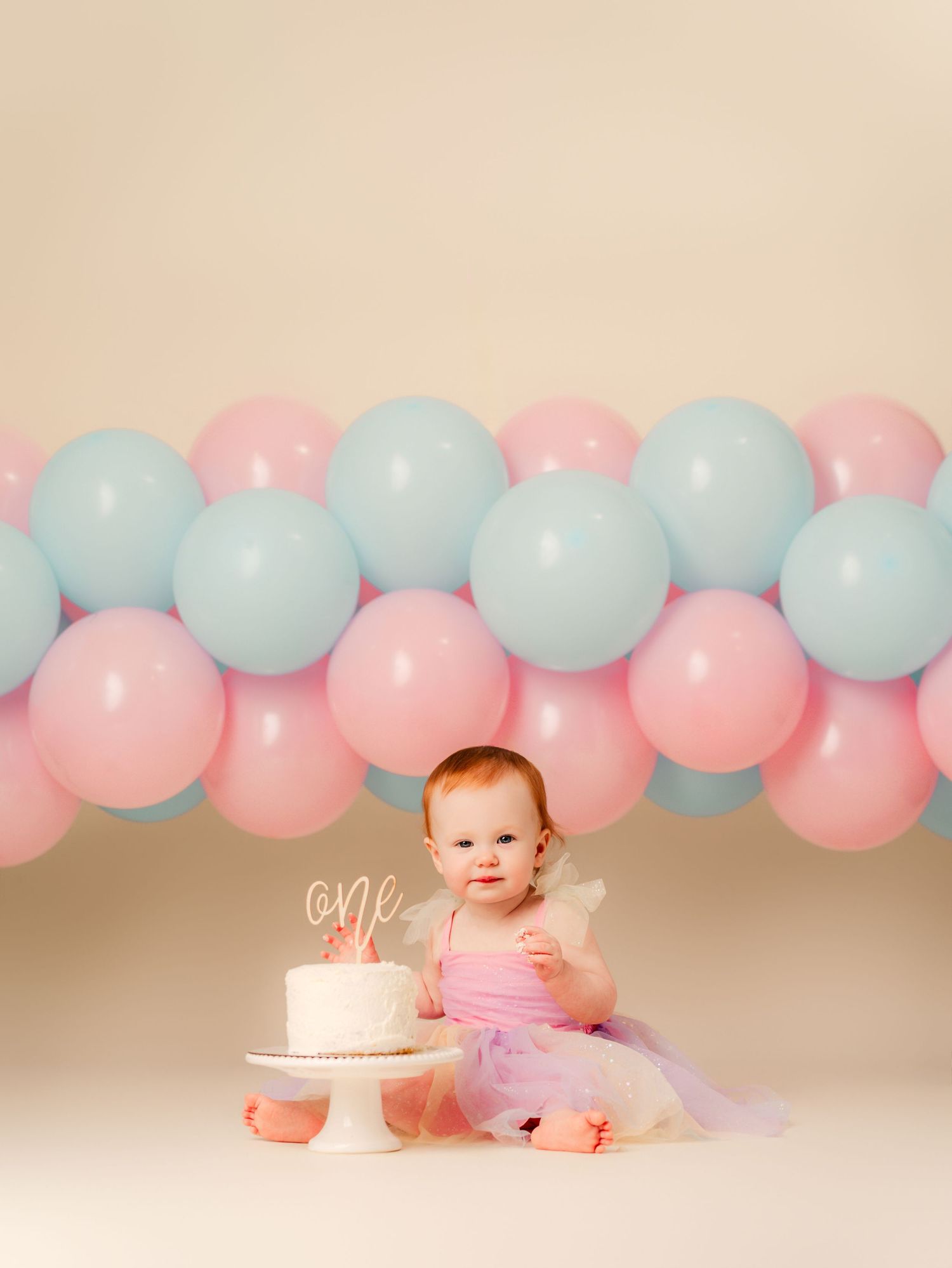 Baby girl in pink tutu sits beside first birthday cake with pink and blue balloon garland backdrop.