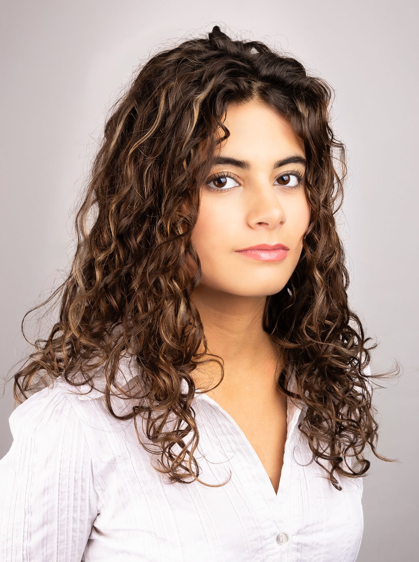 Headshot portrait of a girl in front of a white backdrop, side shot, looking at the camera from the side with a half-smile