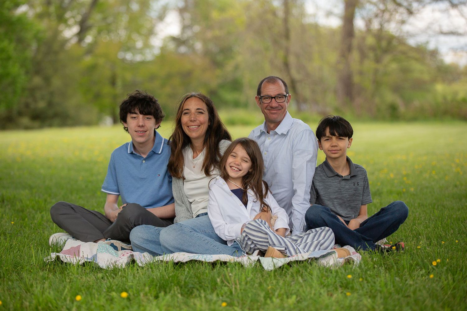 Family of five smiling together on a blanket in a lush green park on a sunny spring day.