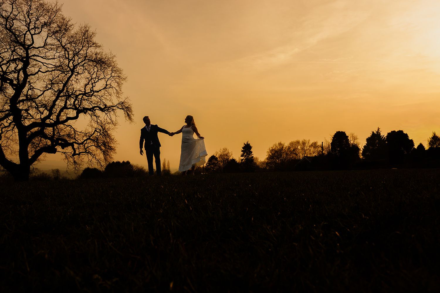 Silhouette of a bride and groom holding hands at sunset in a field with trees against a golden sky. Shireburn Arms