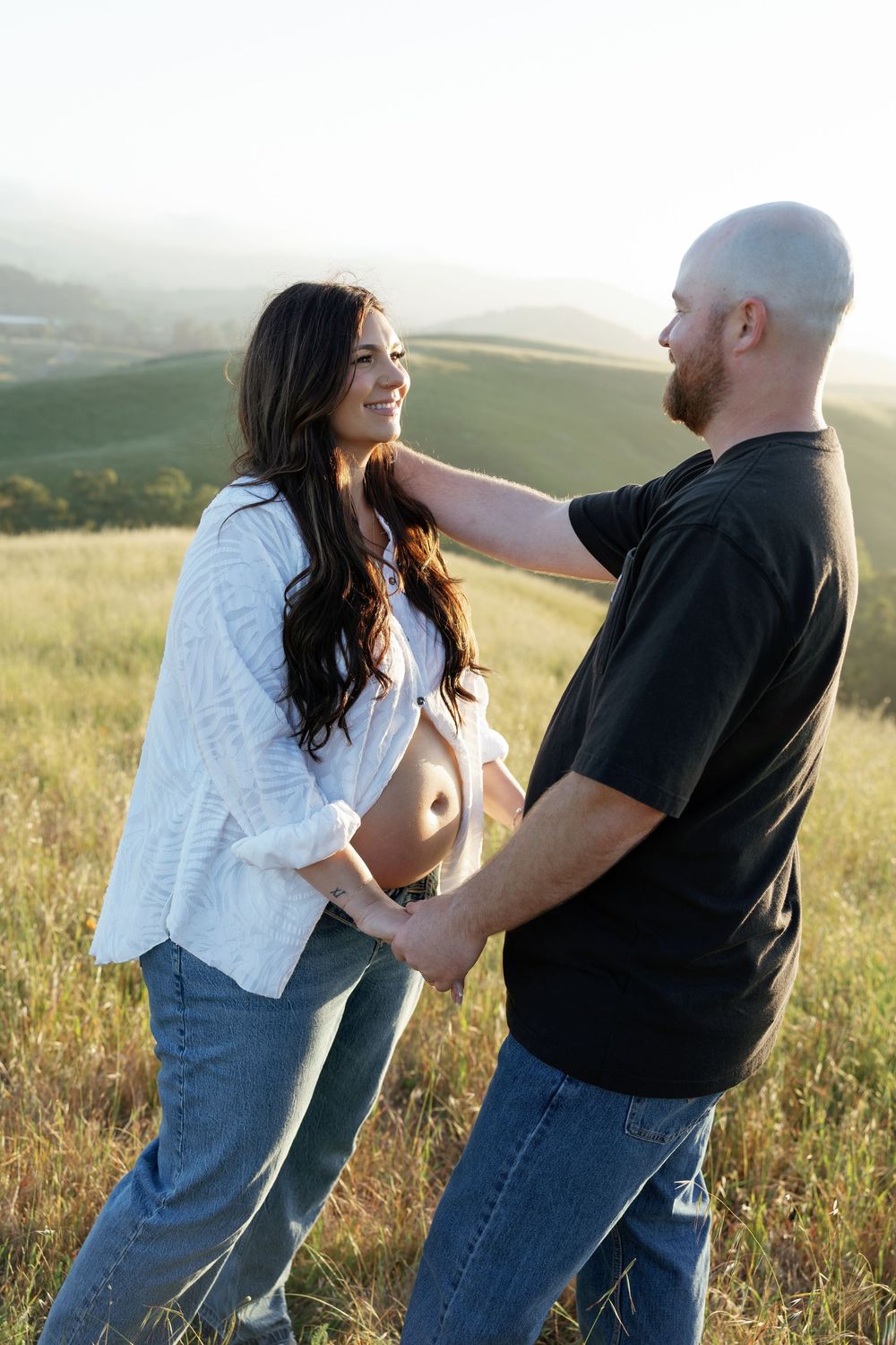 Smiling pregnant woman in white shirt and jeans holds hands with bald man in sunny green hillside field.