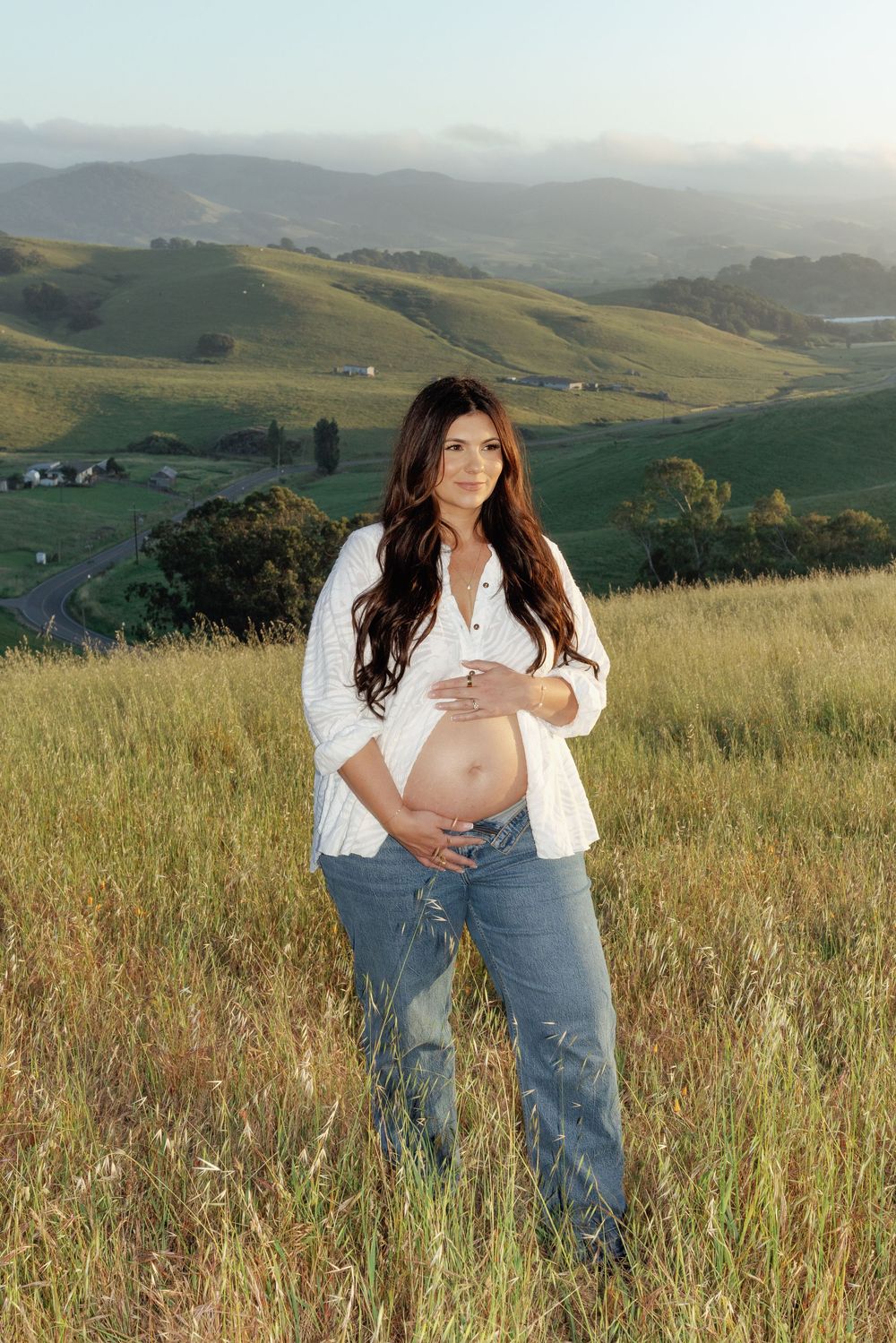 Pregnant woman in white blouse and jeans cradling baby bump in golden field with misty California hills behind her.